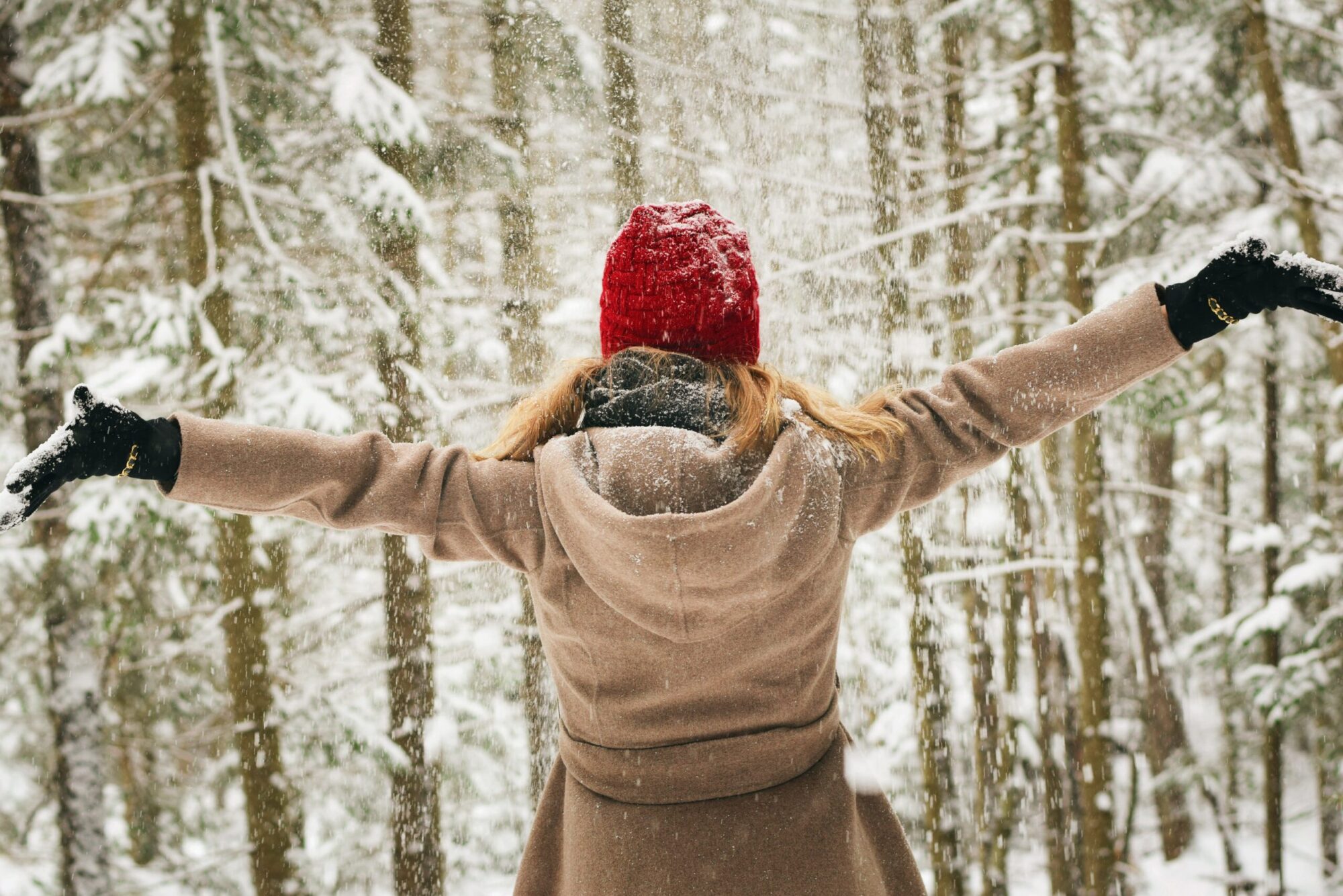 The picure shows a woman wearing a red hat and light brown coat from the back. She has open arms and pictured in a snowy and foresty scene.
