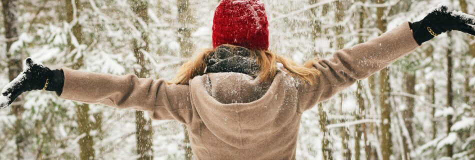 The picure shows a woman wearing a red hat and light brown coat from the back. She has open arms and pictured in a snowy and foresty scene.