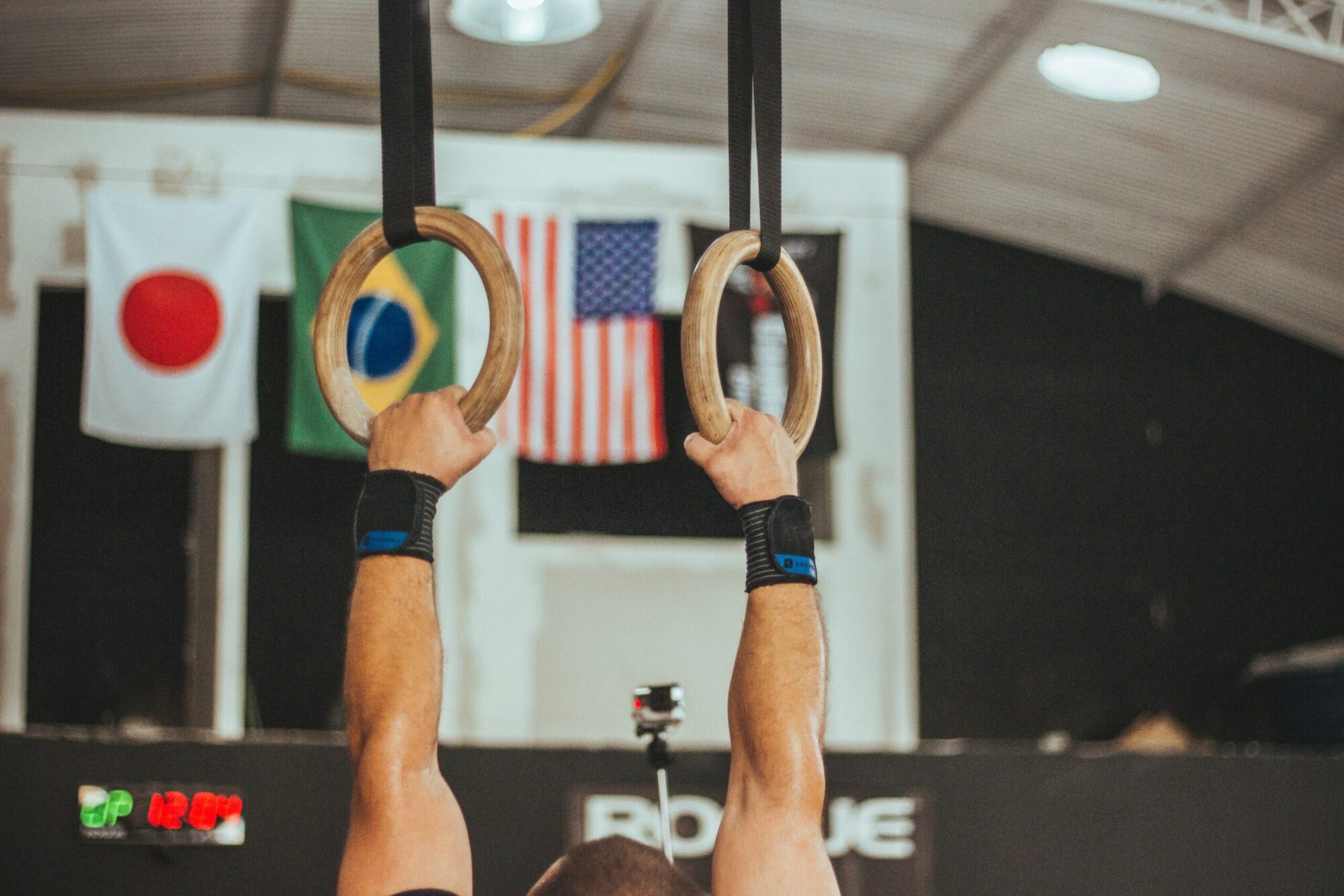 Male gymnast's arms on the rings. And countries flags in the background.